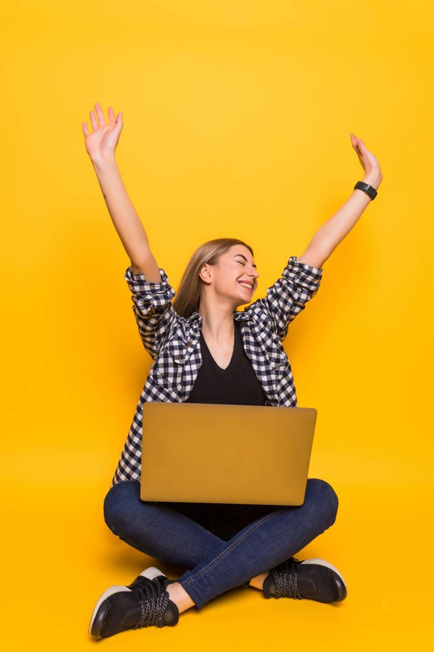 Woman sitting on the floor with a laptop on her lap, raising her hands in excitement