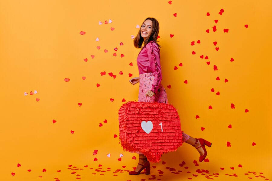 Woman in pink dress holding a red heart-shaped piñata with confetti falling around her